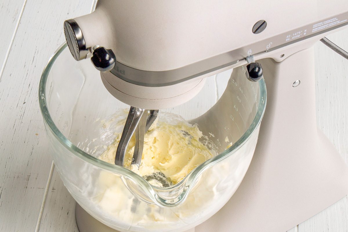 3/4th shot of A KitchenAid stand mixer, in a light gray or off white color, is shown with a clear glass mixing bowl attached; inside the bowl, a pale yellow, creamy mixture, is being stirred by a flat beater attachment; the mixer is positioned on a white wooden surface
