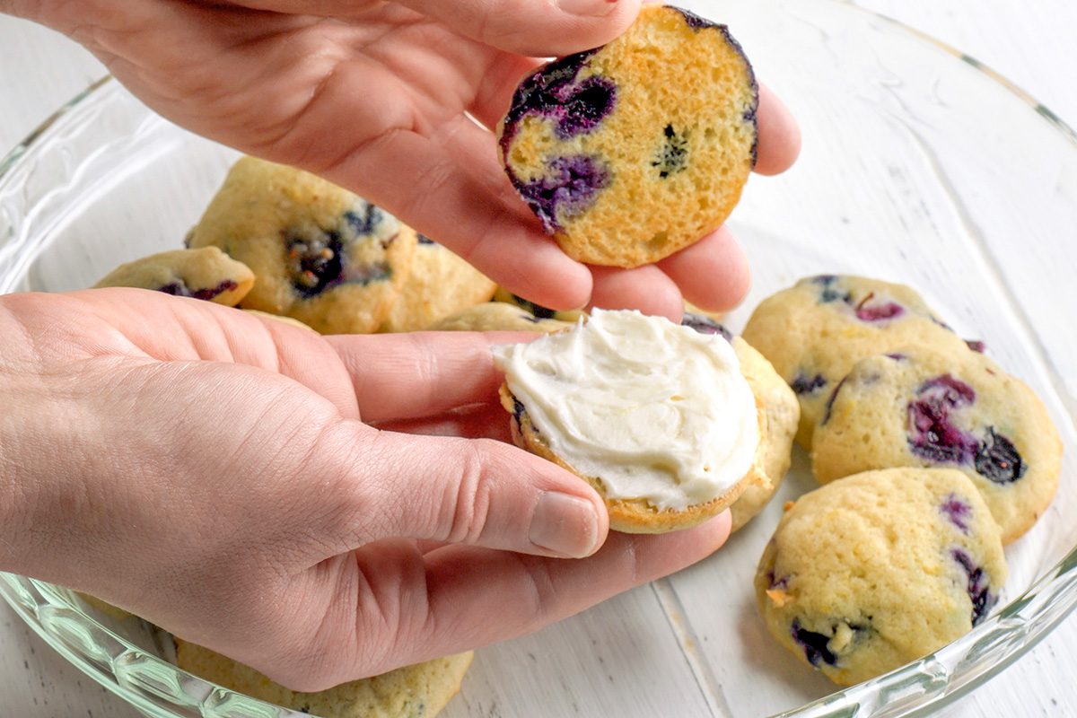 3/4th shot of Two hands are shown holding a blueberry muffin top, and a metal spatula is spreading a thick layer of creamy white frosting on its flat surface; several other similar muffins are visible in a clear glass dish below, the dish rests on a white wooden surface
