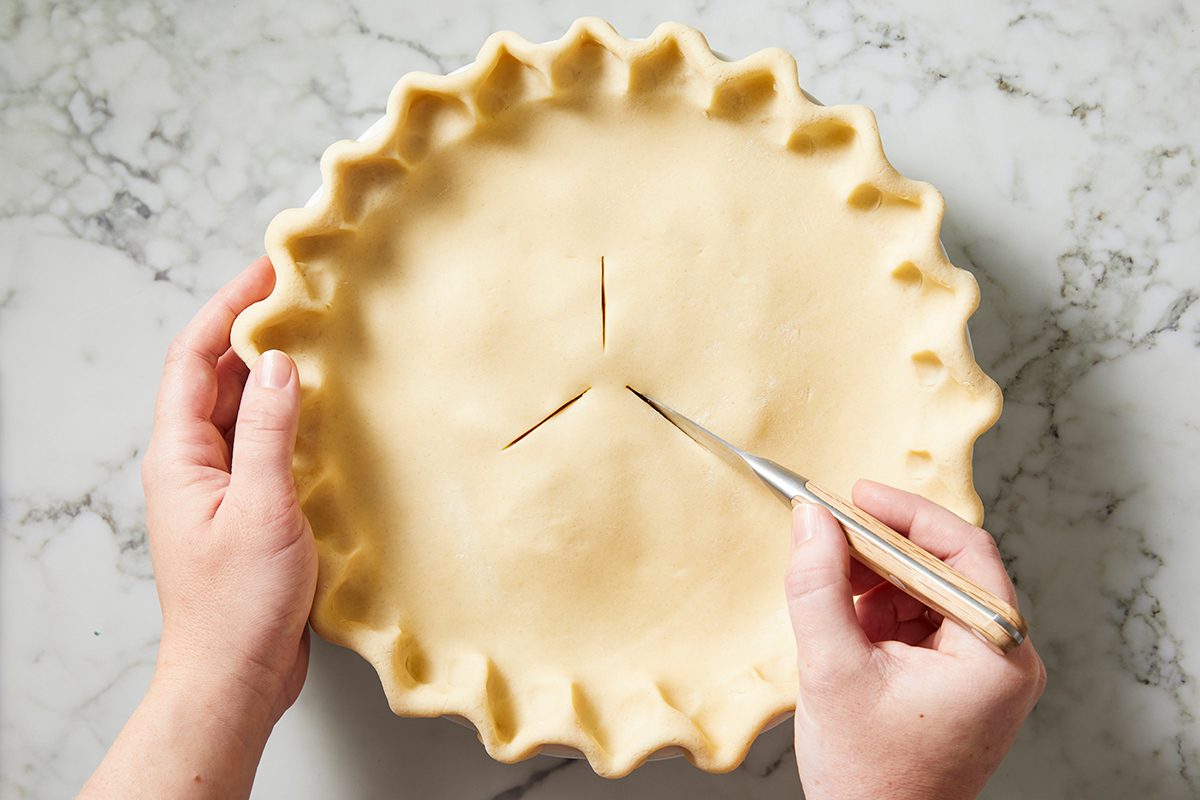 A person holds an unbaked pie with crimped edges on a marble surface, using a knife to cut three slits in the top crust.