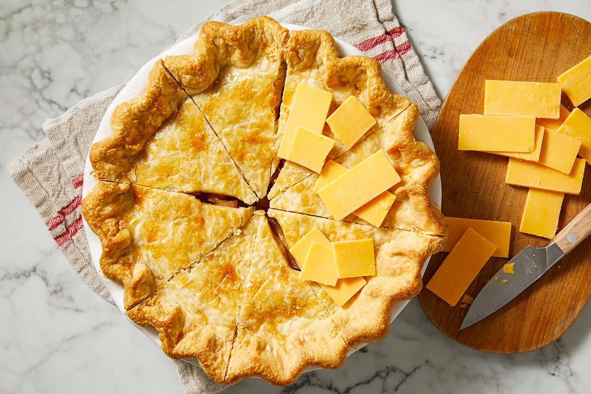 A baked pie with a golden, flaky crust sits on a towel. The pie is sliced, and several slices of cheddar cheese are placed on top and beside it, with a knife and more cheese on a wooden board nearby.