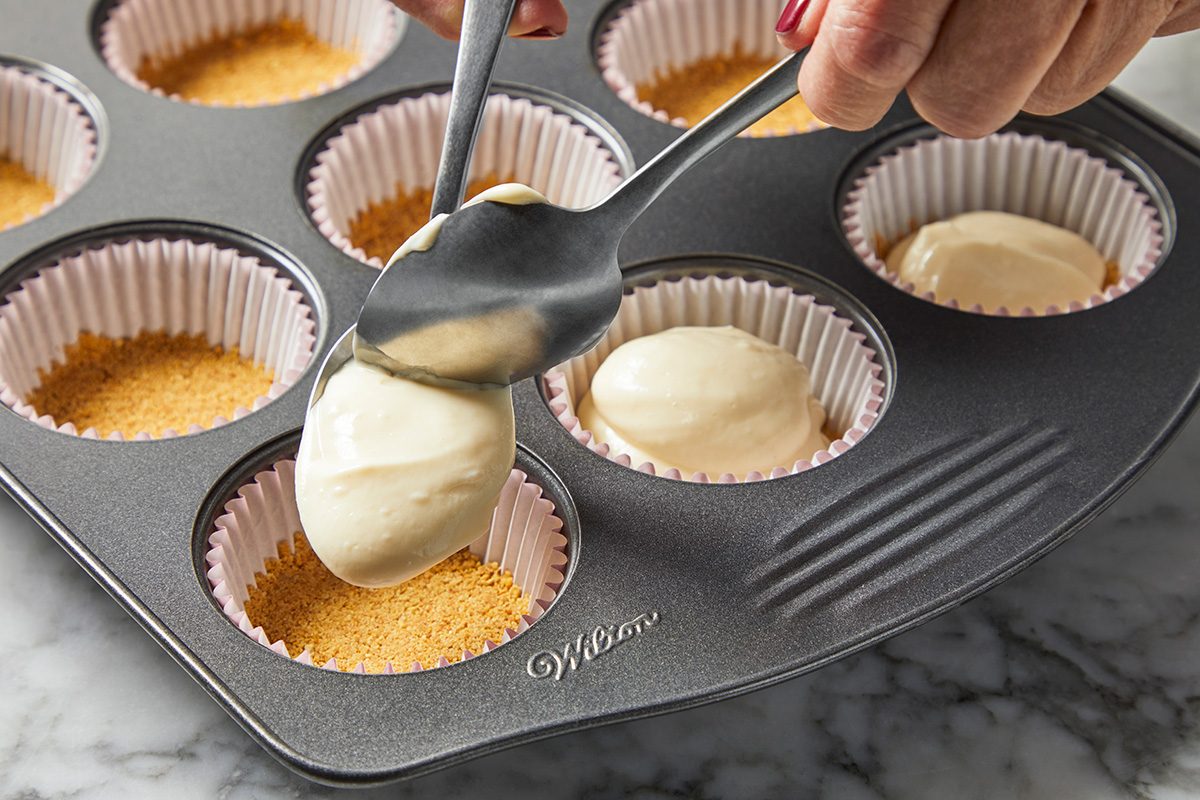 Close-up shot of a hand is seen carefully spooning a creamy mixture into individual cupcake liners placed within a muffin tray; all arrange on a marble surface