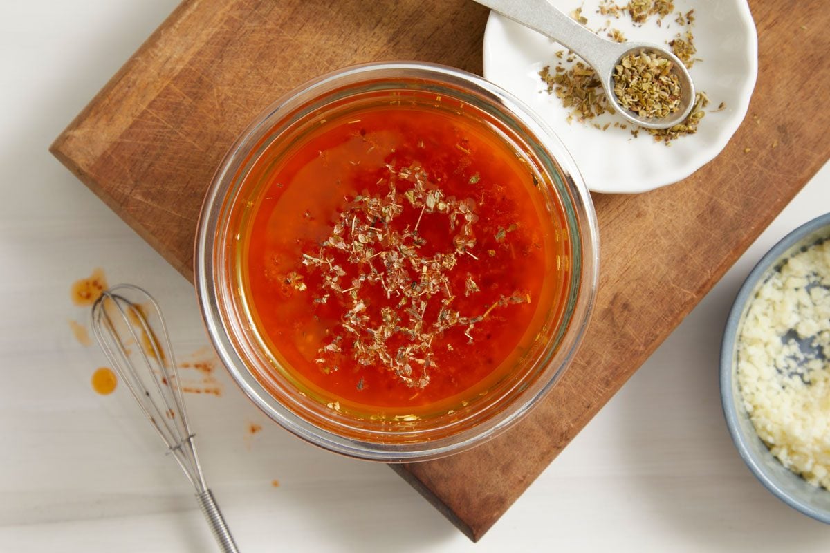 Overhead shot of Sub Oil and Vinegar Drizzle; In a small glass bowl placed over wooden board; whisk together all of the ingredients in bowl; a spoon over a small plate; all set on a cream surface;