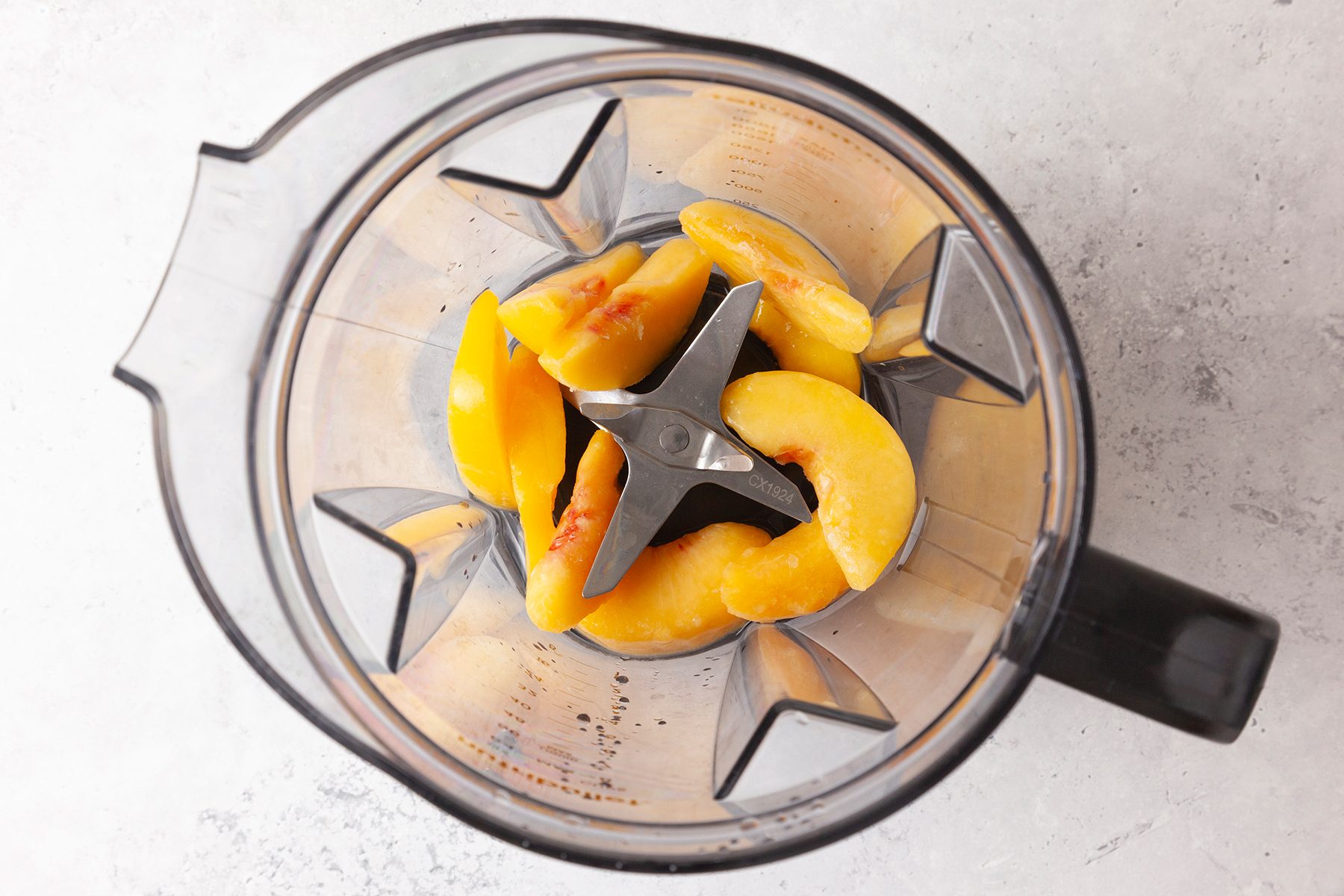 Overhead view of a blender containing several frozen peach slices arranged around the metal blades, on a light-colored surface.