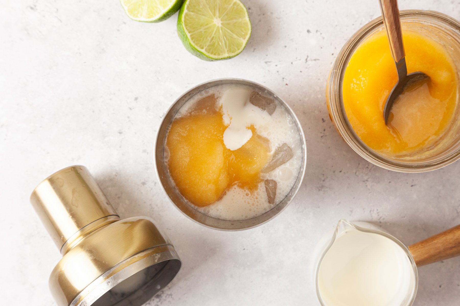 Overhead view of a cocktail shaker with ice, frozen mango, and cream, next to a jar of mango puree with a spoon, a small pitcher of cream, and sliced limes on a light surface.