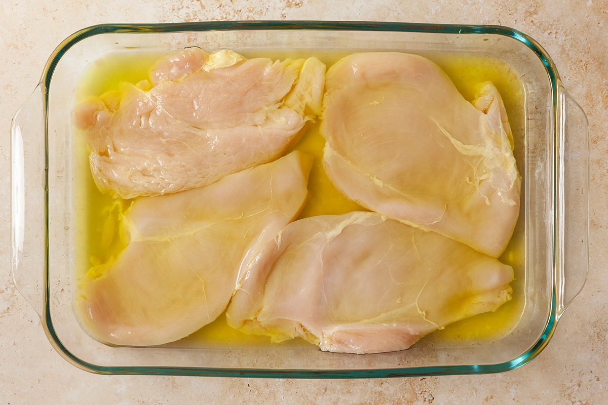 Four raw chicken breasts are placed in a glass baking dish, partially submerged in a yellow liquid, likely oil or marinade, on a light-colored countertop.