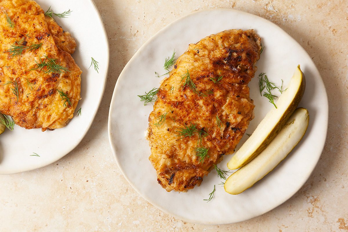 Two plates each hold a piece of breaded, golden-brown fried chicken garnished with fresh herbs, served with two pickle spears on a light-colored surface.