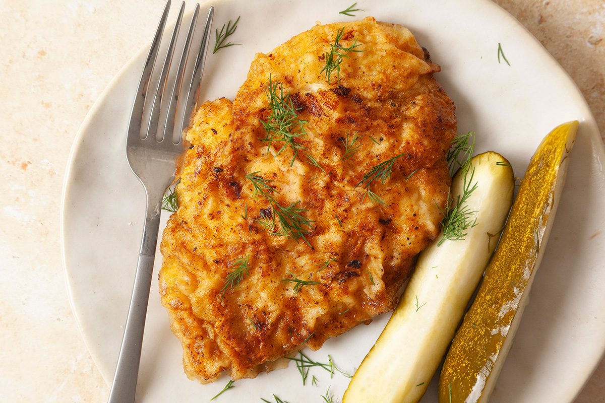 A golden-brown breaded cutlet garnished with dill, served on a white plate alongside two pickle spears, with a fork resting on the plate.