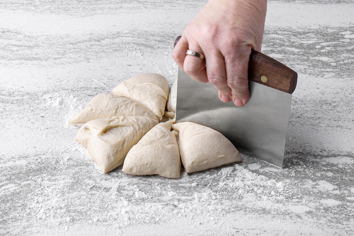 A hand uses a metal bench scraper to cut a round piece of dough into six wedges on a floured surface.