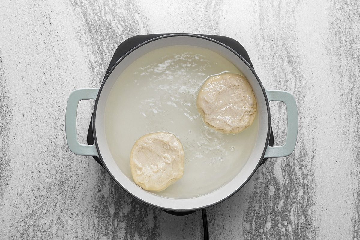 A pot of simmering water on a stovetop with two round pieces of dough cooking inside. The pot has light blue handles and sits on a marbled countertop.