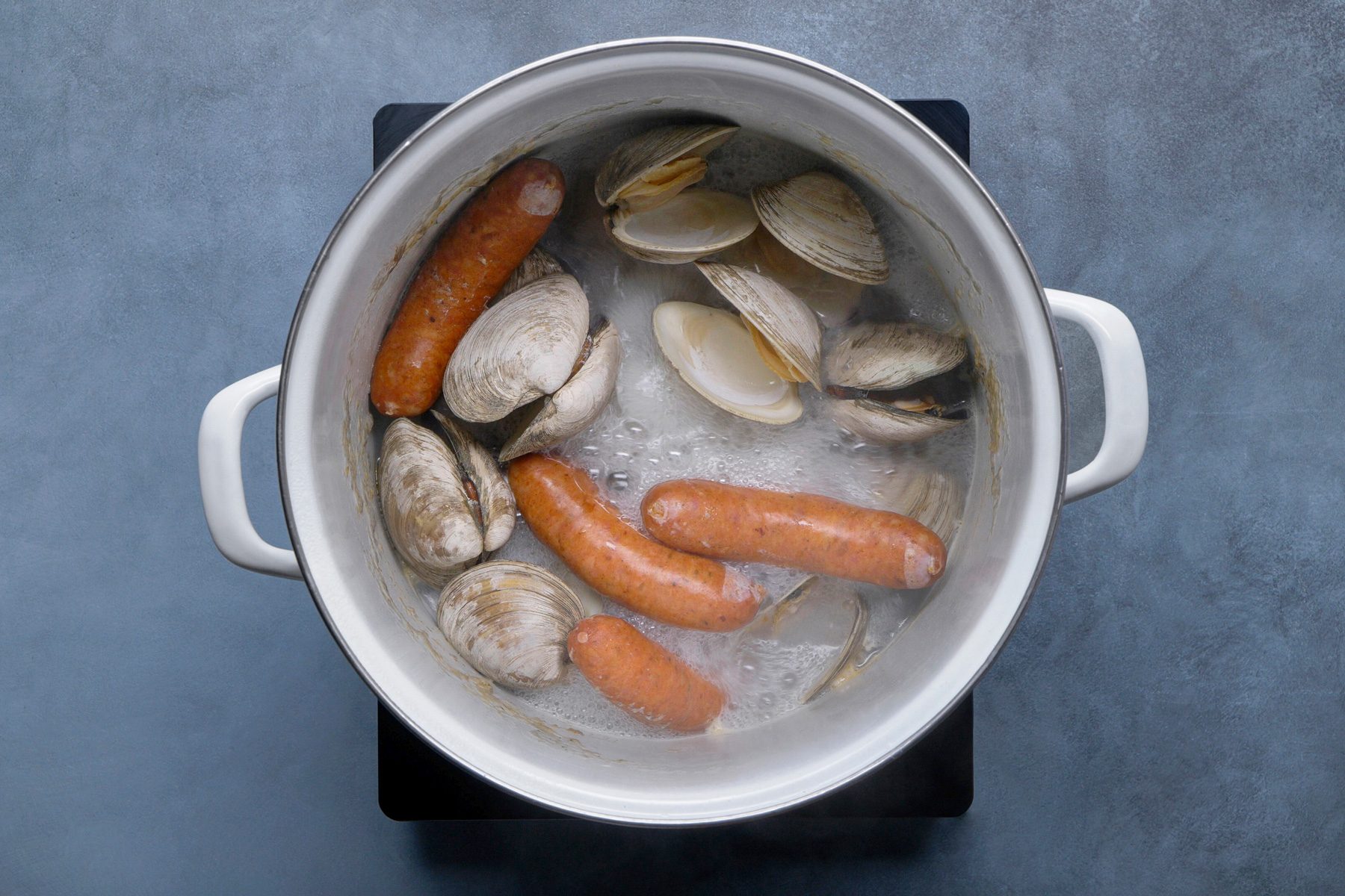 overhead shot of a white pot filled with boiling water, sausages and several open clam shells; the sausages are submerged in the bubbling water, and the clam shells are scattered around them; the pot has white handles and is sitting on a black electric stovetop; the background is a blue gray surface