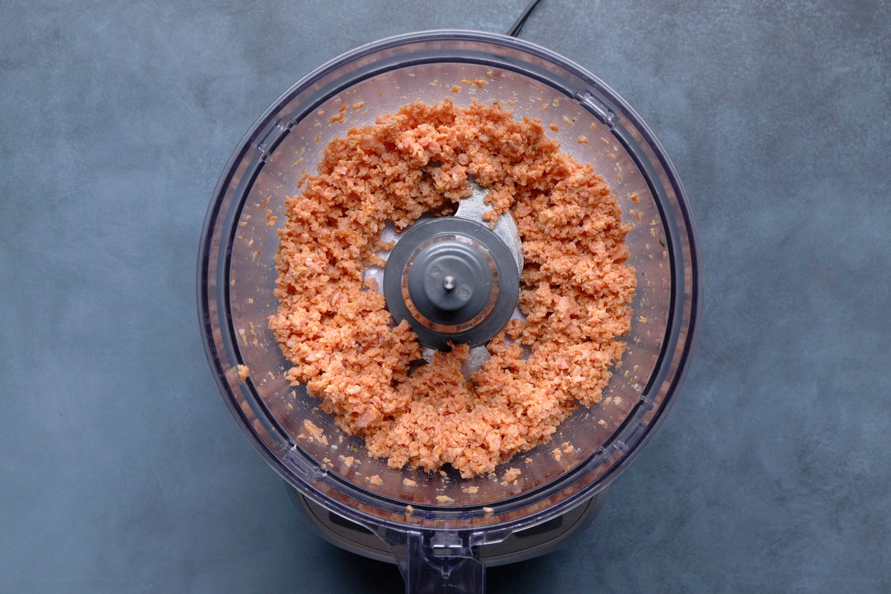 overhead shot of the inside of a clear plastic food processor bowl containing a chunky, light brown mixture, possibly homemade peanut butter or a similar nut butter; the bowl is sitting on the base of the food processor, which is partially visible at the bottom of the frame, and the background is a solid, muted blue gray surface