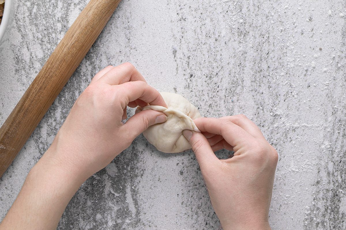 A pair of hands pinches the edges of dough together to seal it, preparing a dumpling on a floured surface. A rolling pin rests nearby.