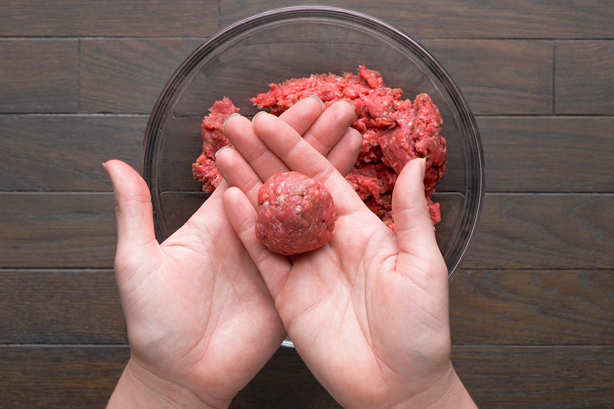 Two hands shape a ball of raw ground meat over a glass bowl filled with more ground meat, on a dark wooden surface.