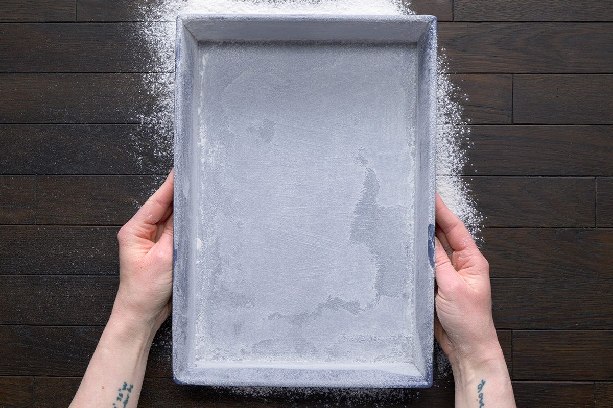 overhead shot of two hands are shown holding a rectangular baking pan over a dark brown wooden surface; dusting flour