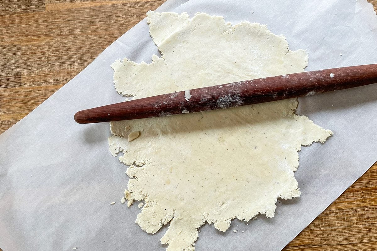 A wooden rolling pin rests on a sheet of parchment paper, rolling out an unevenly shaped piece of dough on a wooden surface.