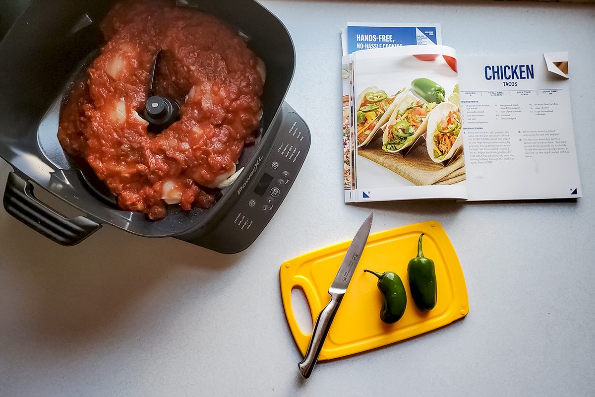 A kitchen counter with a food processor containing ground meat, an open cookbook showing a chicken taco recipe, a yellow cutting board with a knife, and two jalapeños.