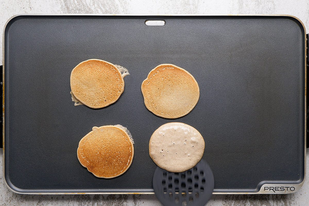 Four pancakes cooking on a rectangular black griddle; three pancakes are flipped, one is being cooked with a black slotted spatula underneath it. The griddle is on a light countertop.