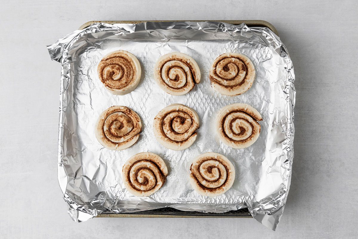 Eight unbaked cinnamon rolls are arranged in three rows on a baking tray lined with crumpled aluminum foil. The cinnamon rolls are evenly spaced on the foil surface.