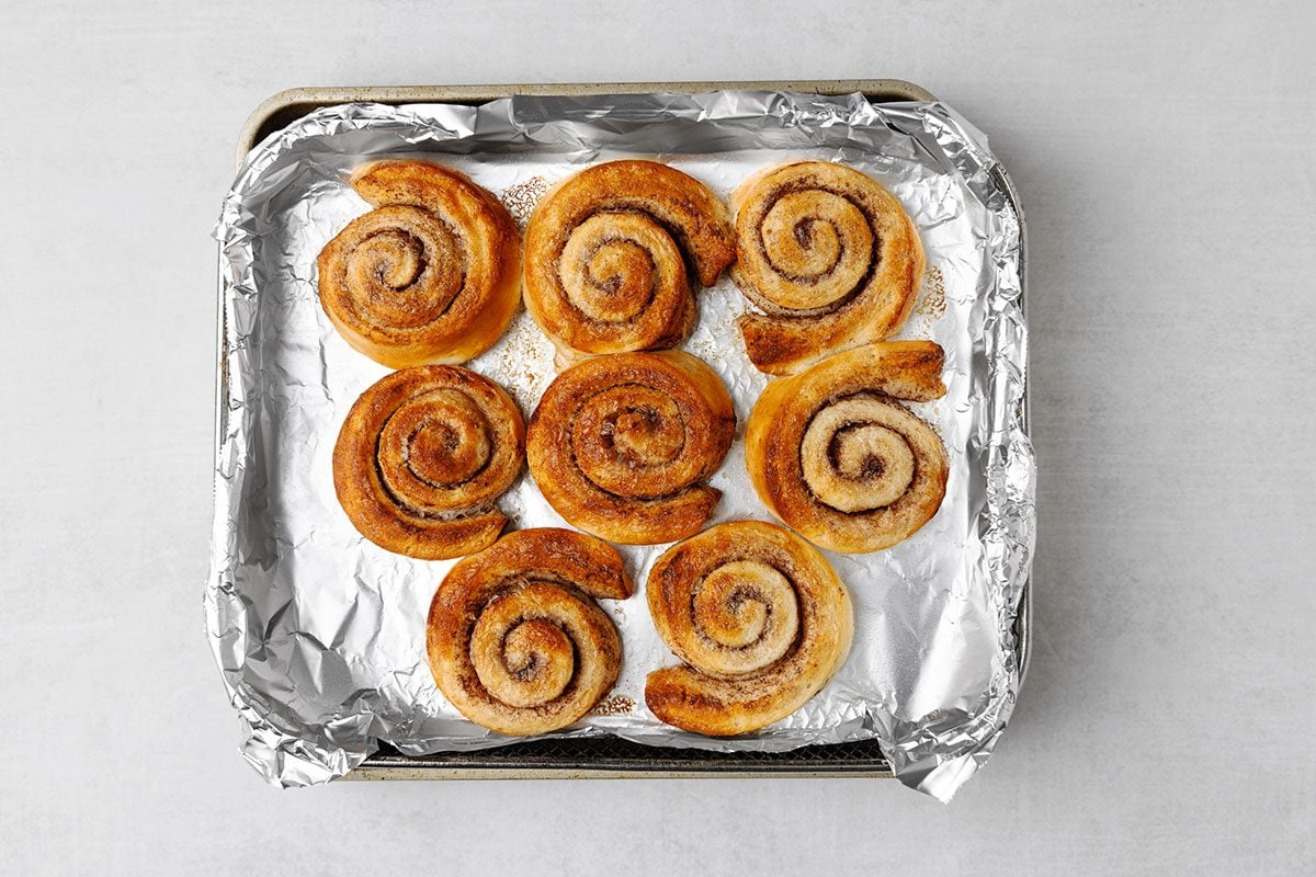 Nine cinnamon rolls arranged on a foil-lined baking tray, viewed from above, with golden brown, spiral-shaped pastry on a light gray background.