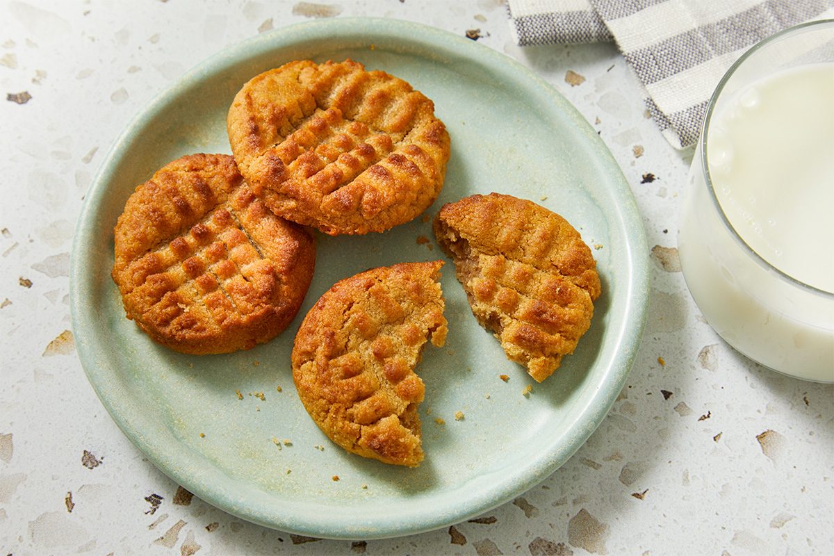 A green plate holds four peanut butter cookies, one of which is broken in half. A glass of milk and a striped napkin are nearby on a speckled countertop.