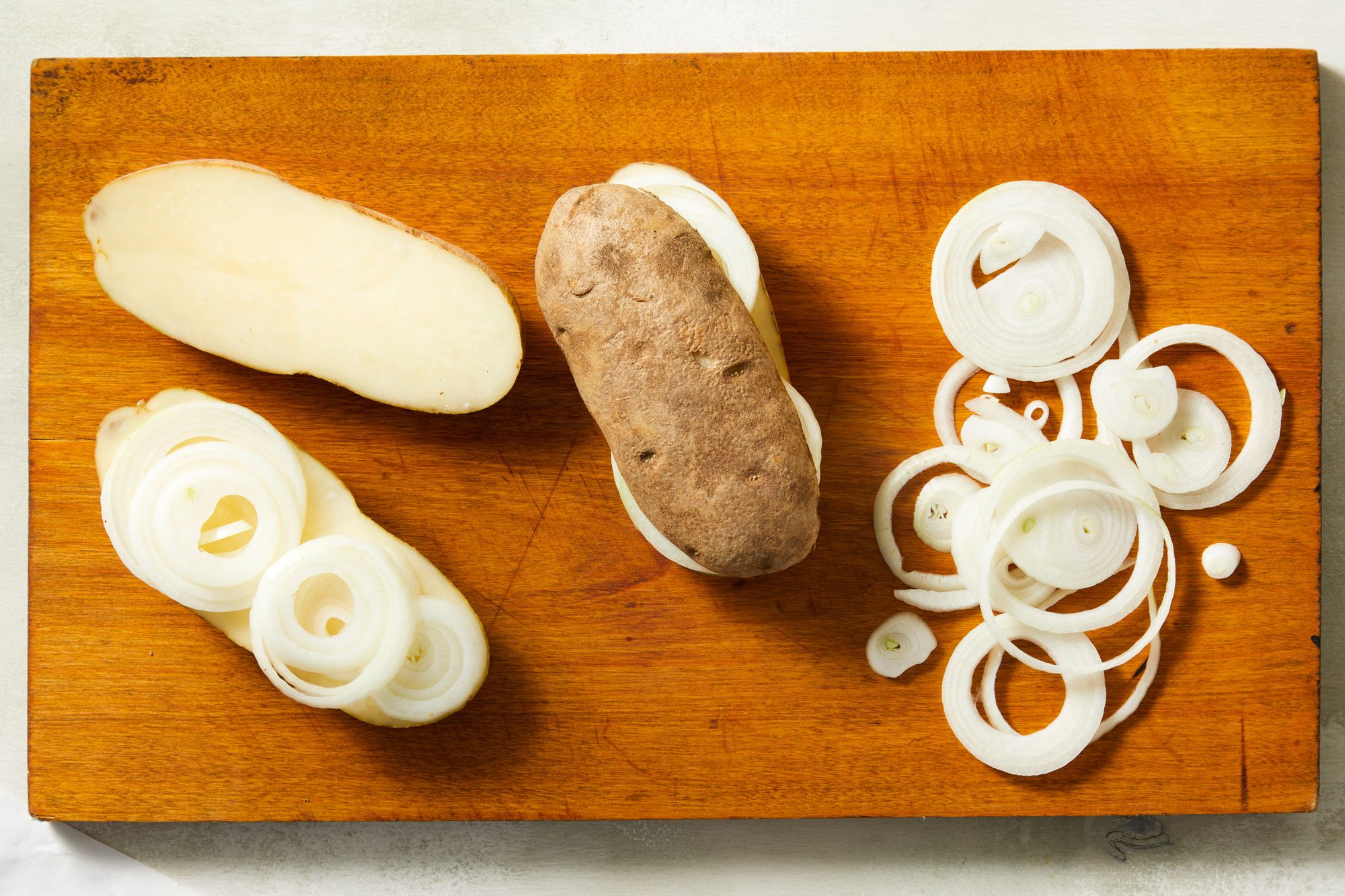 Overhead shot of layer onion slices on cut side of two potato halves; top with other potato half; wooden chopping board; on marble surface