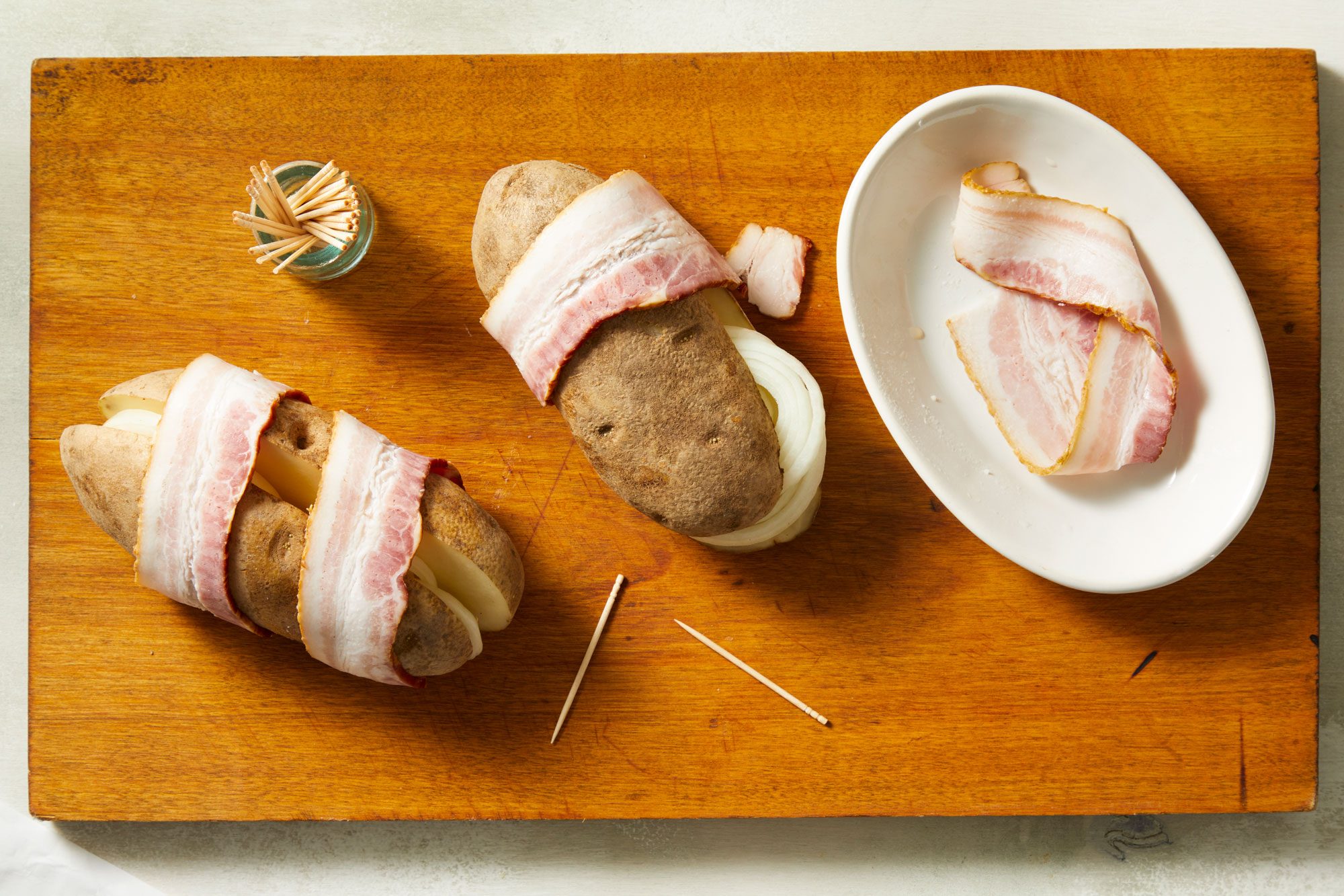 Overhead shot of wrap each potato with two bacon strips on wooden chopping board; Secure with toothpicks; all set on a marble surface
