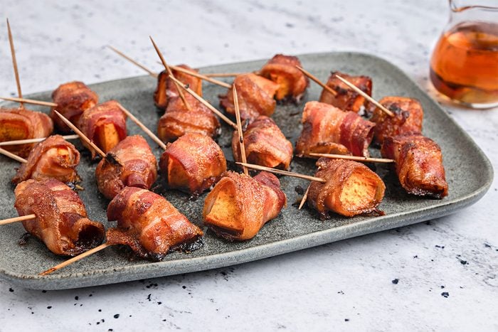 A gray rectangular plate holds several bacon-wrapped sweet potato bites secured with toothpicks. A small glass container of syrup sits in the background on a light, speckled surface.