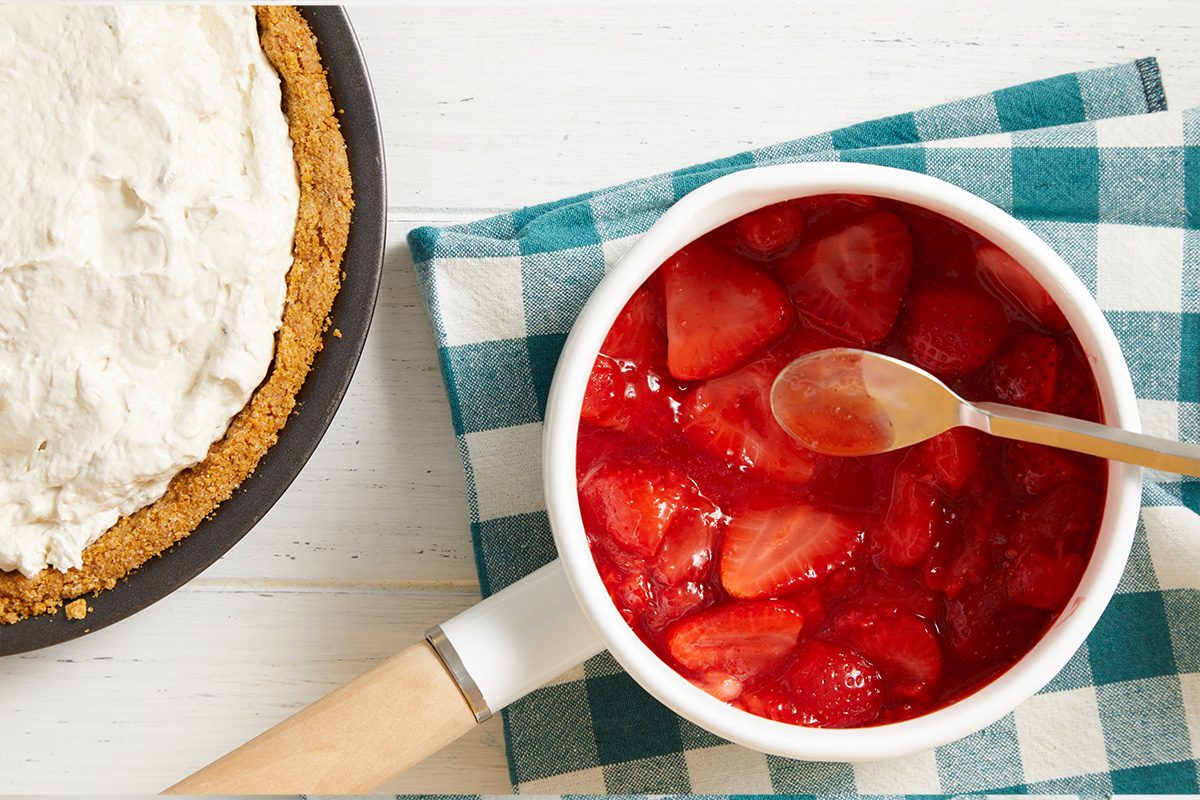 A saucepan of strawberry topping with a spoon rests on a blue and white checkered cloth next to a partially visible pie with a graham cracker crust and whipped cream topping.