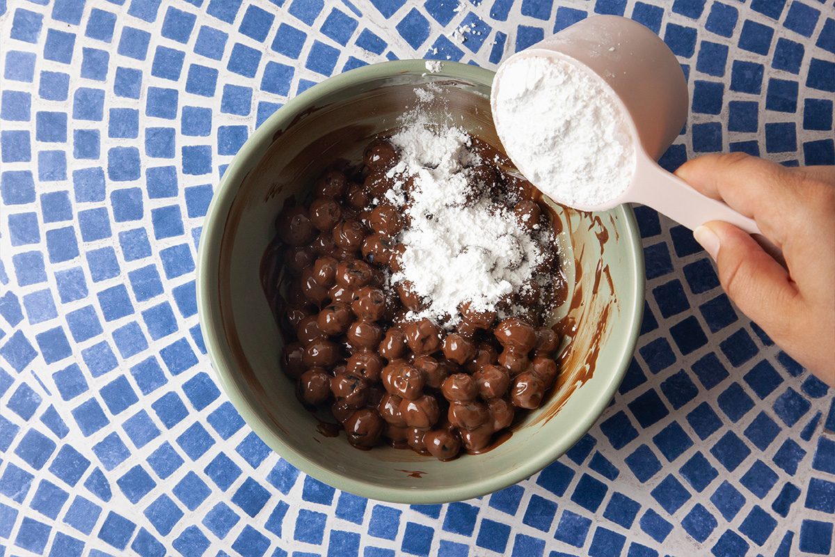 A hand pours powdered sugar from a measuring cup into a bowl of chocolate-coated cereal balls, set on a blue and white mosaic surface.