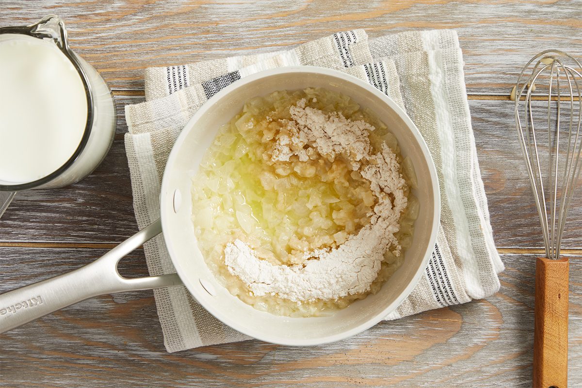 A saucepan with sautéed onions and flour sits on a striped towel. A glass pitcher of milk and a whisk are nearby on a wooden surface.
