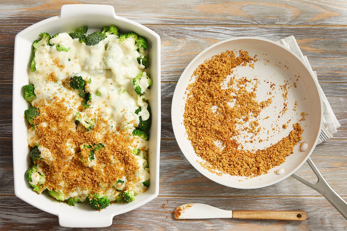 A casserole dish filled with broccoli and creamy sauce, topped with golden breadcrumbs, sits next to a white skillet containing leftover toasted breadcrumbs on a wooden surface.