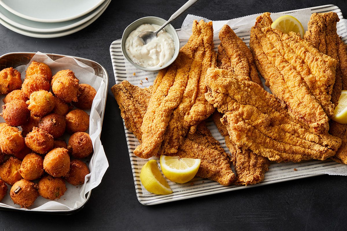 Platter of golden fried fish fillets with lemon wedges and tartar sauce, next to a tray of round hush puppies on parchment paper, with a stack of white plates in the background.