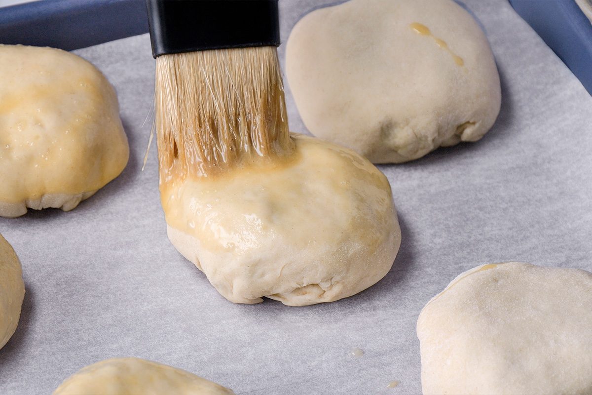 A pastry brush is being used to apply an egg wash to unbaked dough balls on a parchment-lined baking sheet.