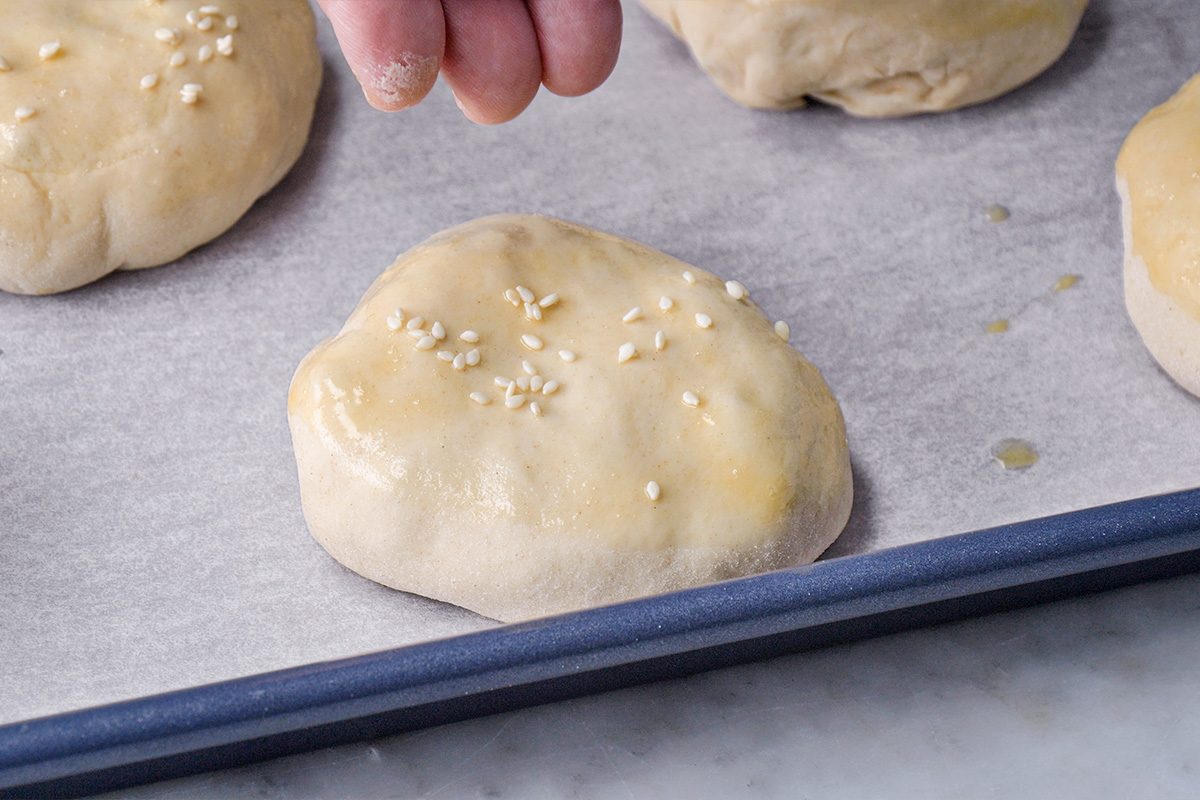 A hand sprinkles sesame seeds onto a round, unbaked dough bun resting on a parchment-lined baking sheet with other dough buns nearby.