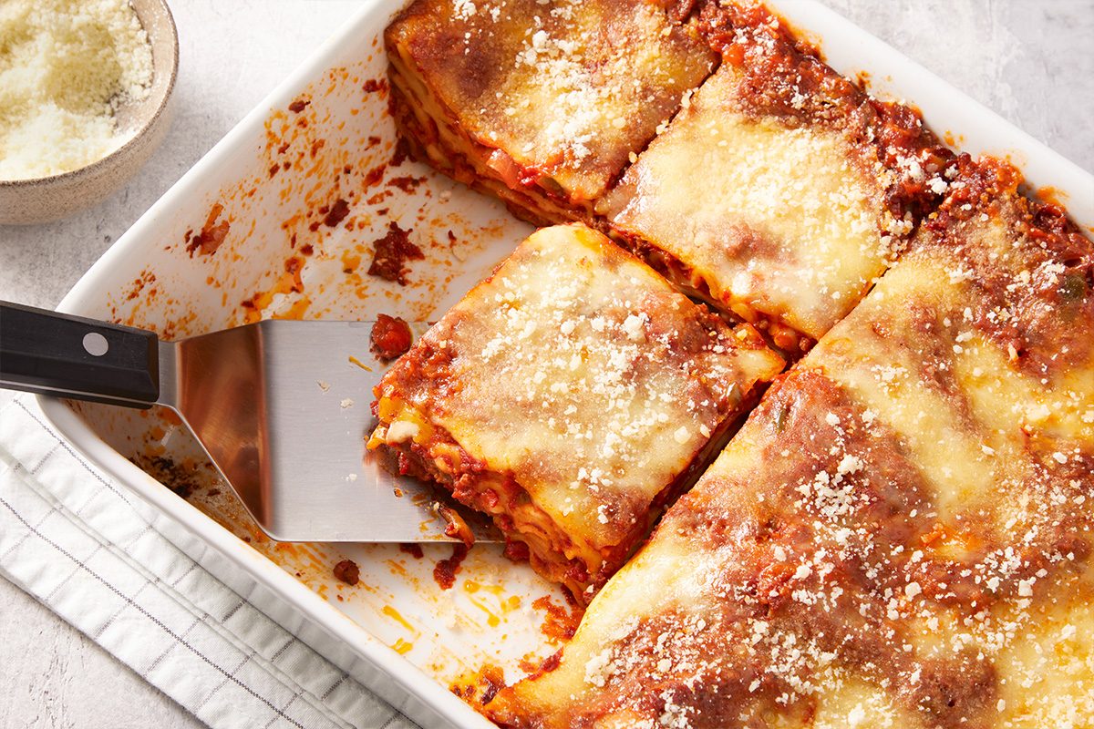 A baking dish filled with baked lasagna, with one cheesy slice being lifted out by a spatula. The lasagna has visible layers of pasta, sauce, and melted cheese on top.