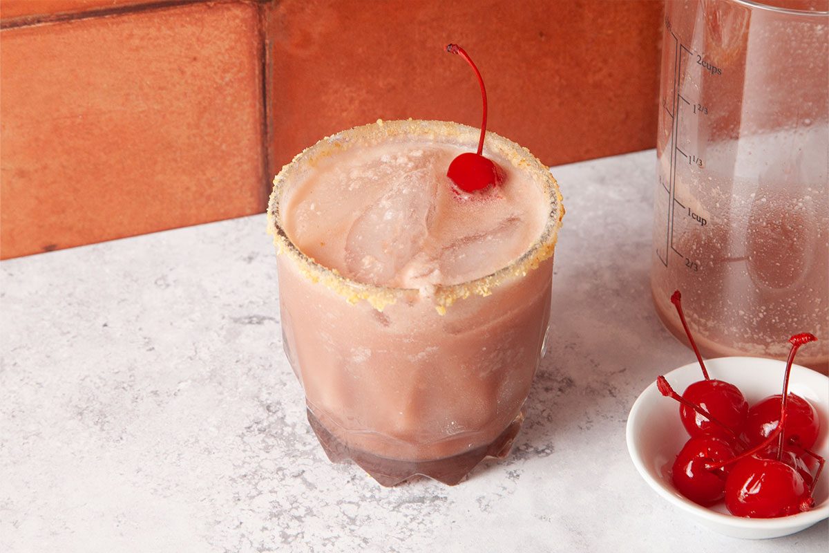 A creamy pink cocktail in a glass with a sugared rim, garnished with a cherry on top. Next to it is a small bowl containing several maraschino cherries, all set against a light countertop and brick background.
