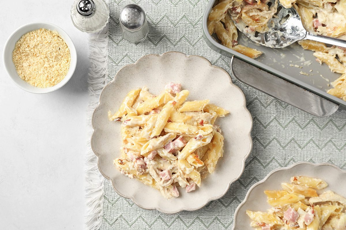 A plate of baked penne pasta with cheese and ham, served on a scalloped-edge white plate. A casserole dish with more pasta, a bowl of grated cheese, and salt and pepper shakers are nearby on a patterned tablecloth.
