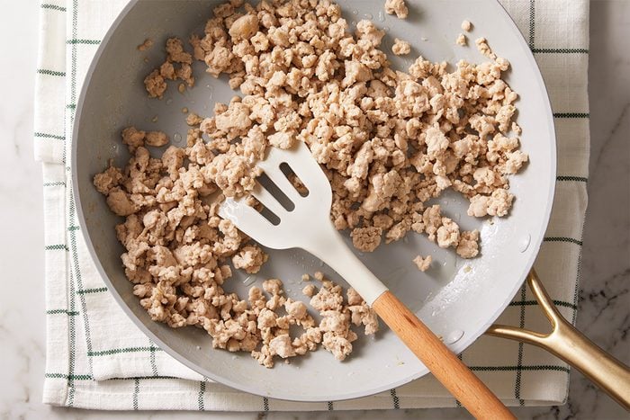 A white frying pan with cooked ground turkey and a white slotted spatula, resting on a white and green checkered kitchen towel on a marble countertop.