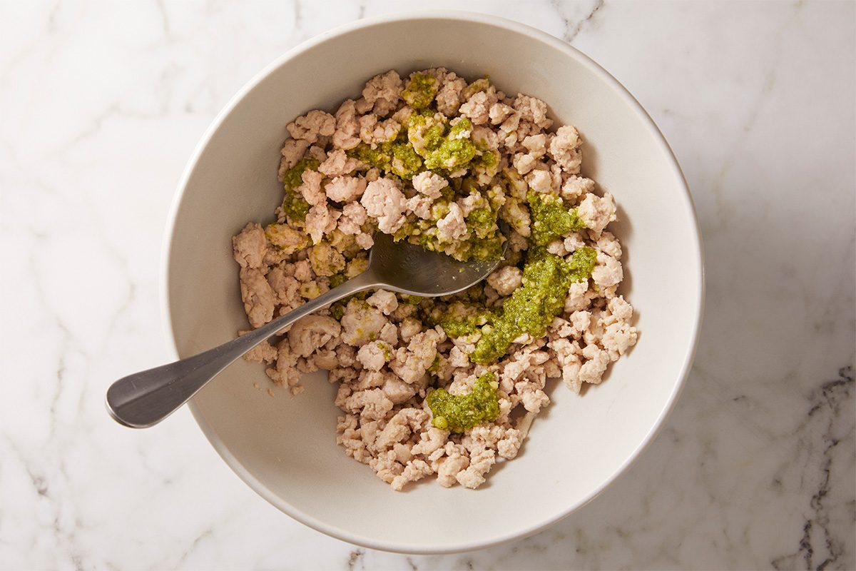 A bowl filled with cooked ground meat and green seasoning, with a spoon resting inside, placed on a white marble surface.