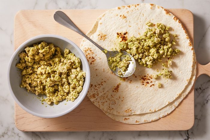 A bowl of tofu scramble with a spoon rests next to a tortilla on a wooden cutting board. Some tofu scramble is being placed onto the tortilla, ready to be wrapped.