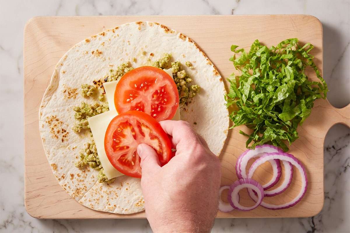 A hand places tomato slices on a tortilla with cheese and mashed avocado on a wooden board, next to chopped lettuce and red onion slices.