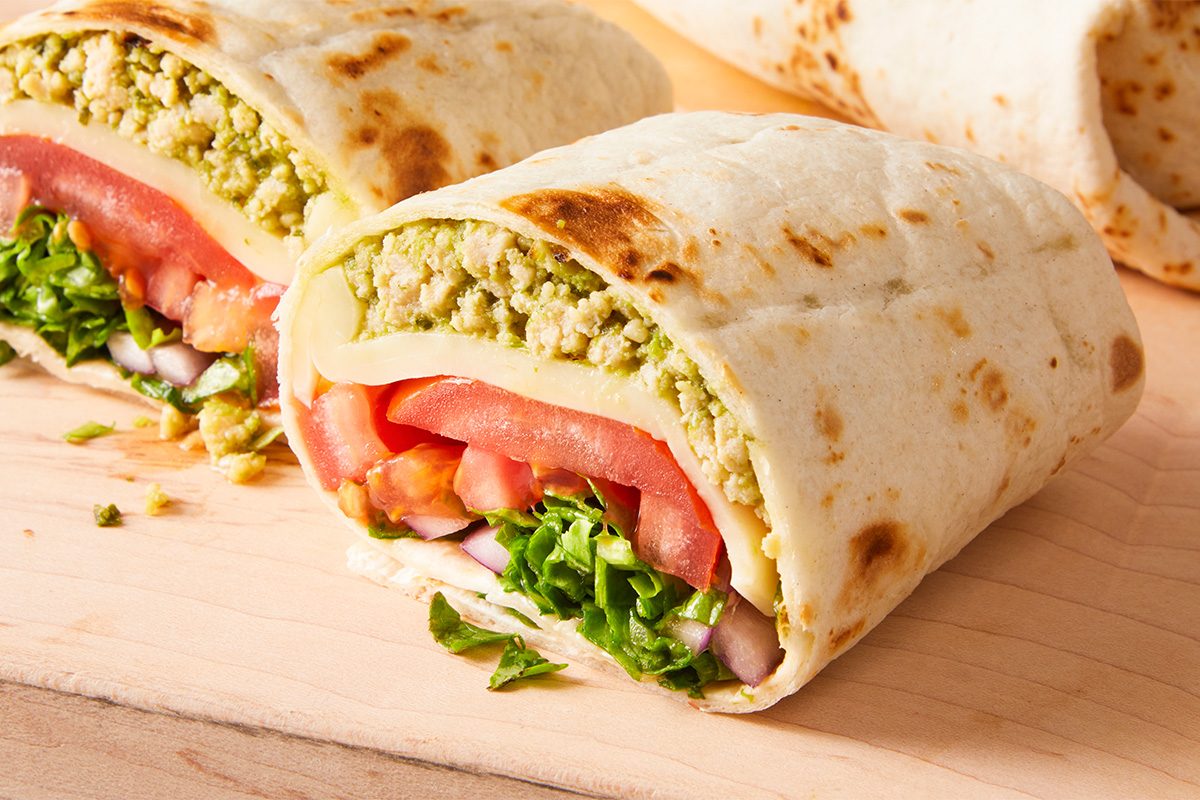 A close-up of a sliced wrap filled with lettuce, tomato, onion, cheese, guacamole, and possibly a grain or tofu, all inside a flour tortilla on a wooden surface.