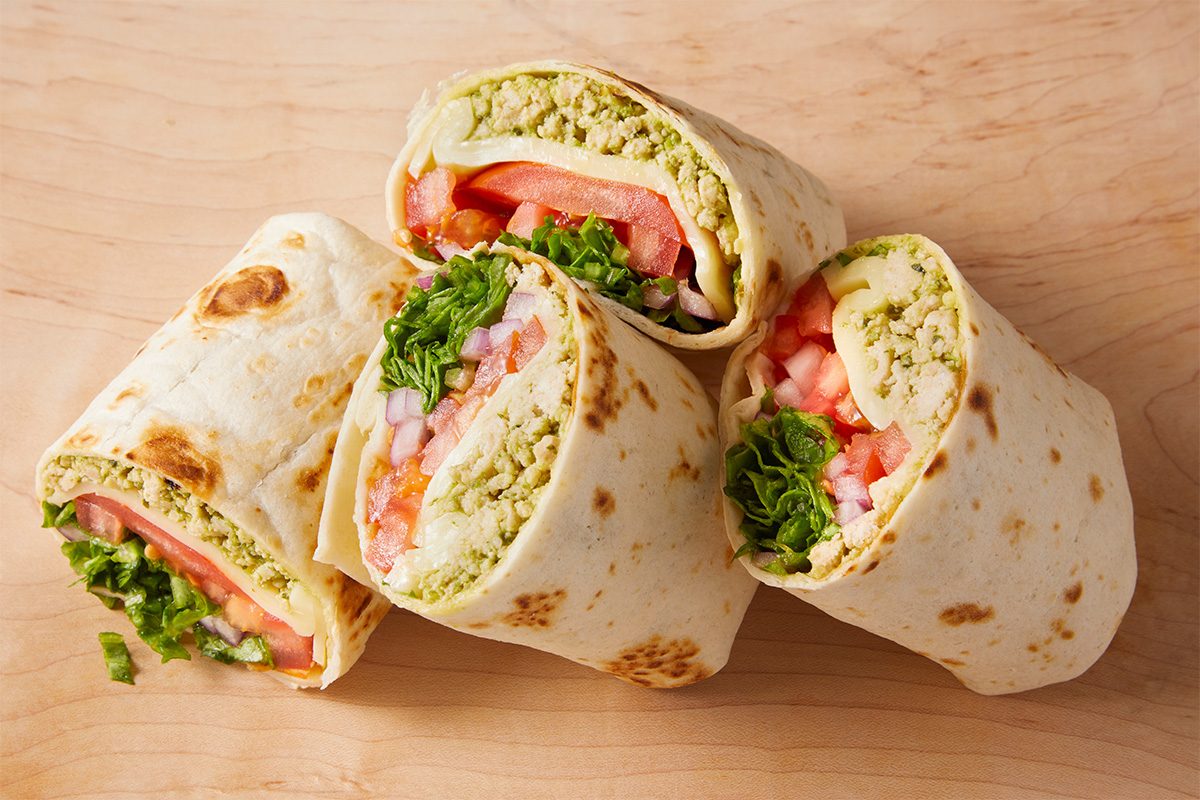 Four tortilla wraps, each cut in half, showing fillings of chopped lettuce, tomato slices, diced red onion, and a crumbly green spread, arranged on a wooden surface.
