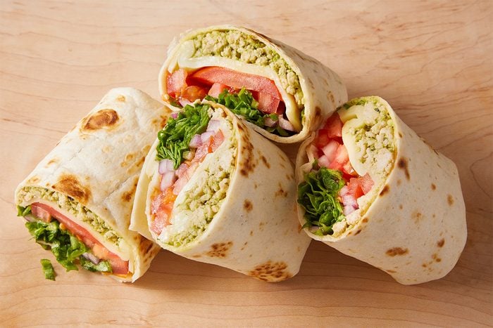 Four tortilla wraps, each cut in half, showing fillings of chopped lettuce, tomato slices, diced red onion, and a crumbly green spread, arranged on a wooden surface.