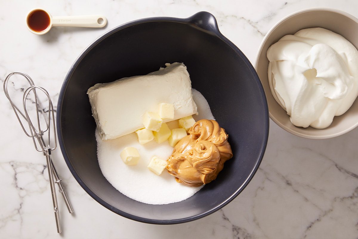 A black mixing bowl with cream cheese, butter cubes, sugar, and peanut butter on a marble countertop. Nearby are a bowl of whipped cream, a hand mixer, measuring spoon with vanilla, and a whisk attachment.