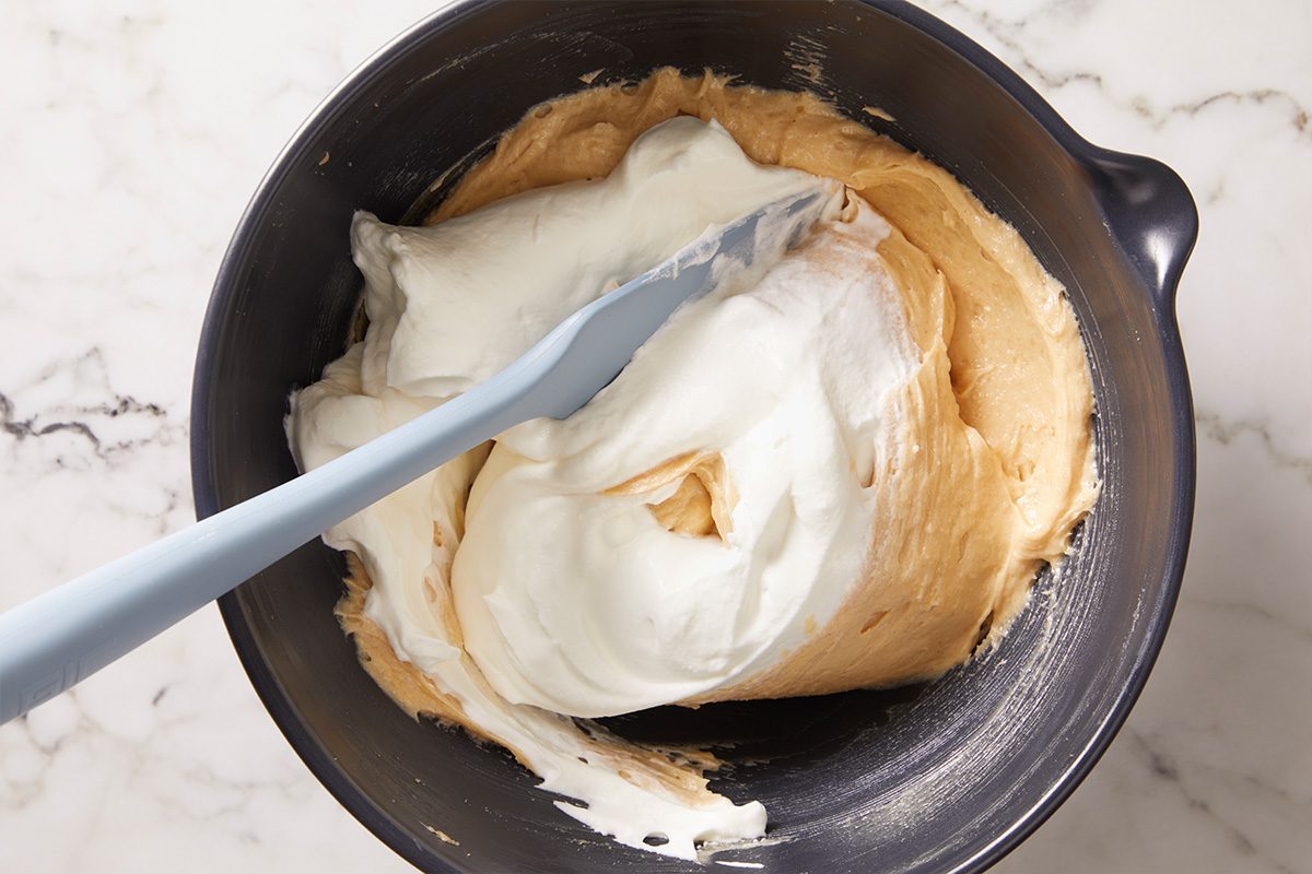 A mixing bowl with a pale, creamy batter being mixed with fluffy egg whites using a light blue spatula on a marble countertop.