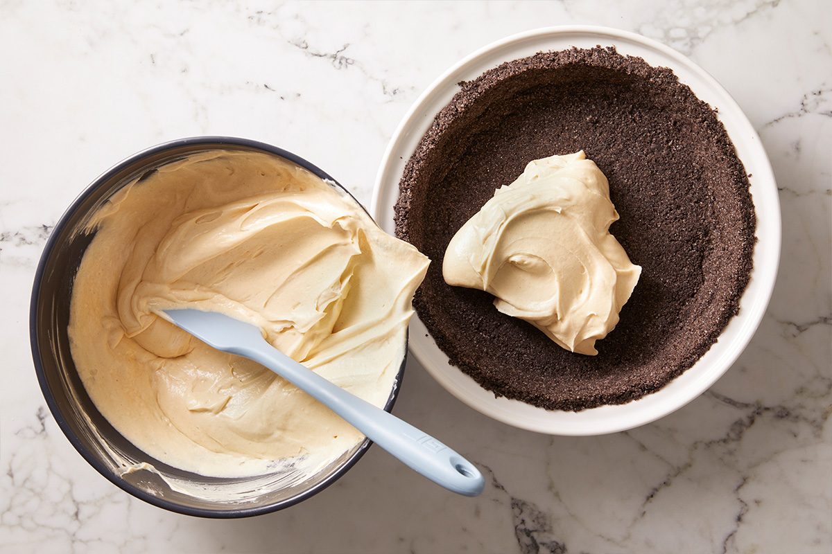 A bowl of creamy filling with a spatula beside a pie dish containing a chocolate crumb crust and a dollop of filling on a marble countertop.