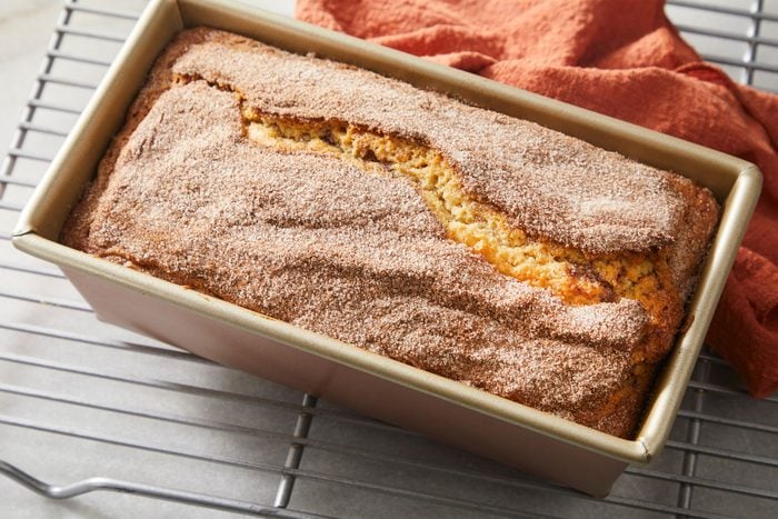 overhead shot of a freshly baked cinnamon sugar bread loaf sits in a rectangular metal pan, resting on a cooling rack with an orange cloth nearby, The loaf has a cracked top and a generous dusting of cinnamon sugar