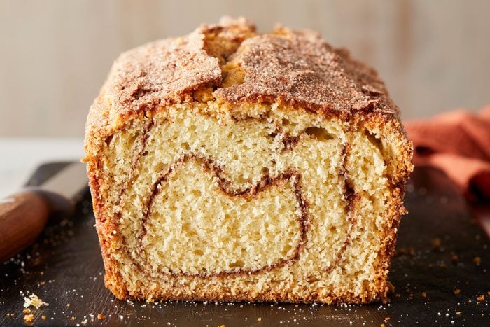 A closeup shot of a sliced loaf of cinnamon swirl bread on a wooden cutting board, showing a spiral pattern inside and a crispy, sugared crust on top