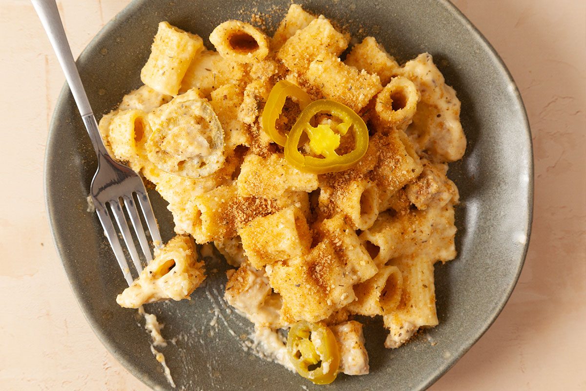 A plate of baked macaroni and cheese with rigatoni pasta, topped with breadcrumbs and sliced jalapeños, sits on a gray dish with a fork.